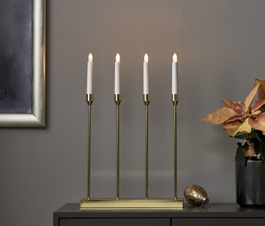 Goldener Kerzenständer mit vier leuchtenden LED-Kerzen auf dunklem Schrank, daneben orangefarbener Weihnachtsstern und goldene Dekoration. Im Hintergrund ein silberner Bilderrahmen.