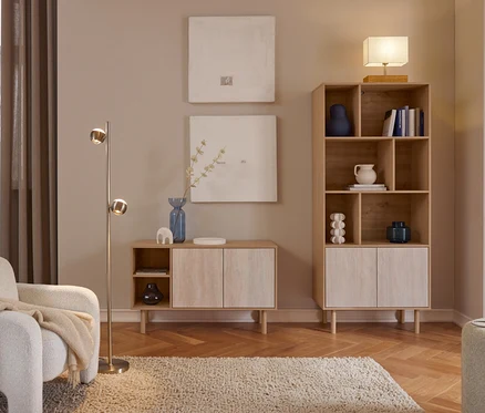 Gemütliches Wohnzimmer mit hellen Holzmöbeln. Ein weißer Sessel mit beiger Decke steht neben einer Messing-Stehlampe. Zwei abstrakte Bilder hängen über einem niedrigen Schrank mit blauer Vase. Ein hohes Bücherregal mit Deko und Büchern wird von einer Tischlampe gekrönt. Ein zotteliger Teppich liegt auf einem Fischgrätparkett.