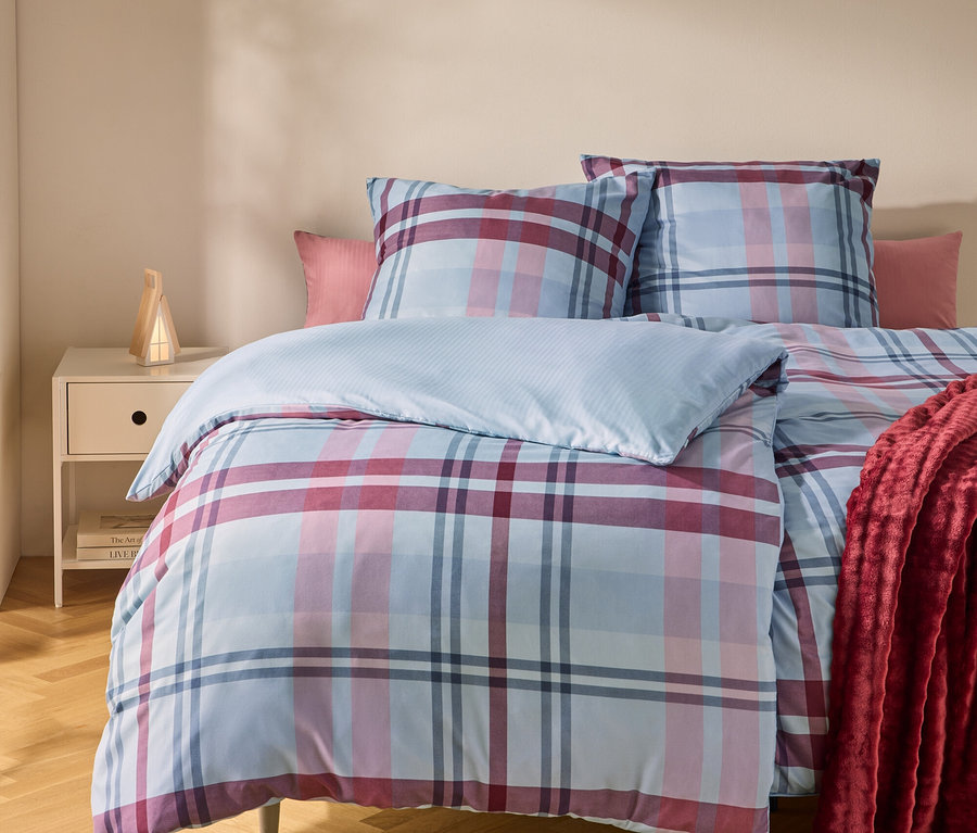 Ein Bett mit einer blau, rosa und rot karierten Bettdecke und Kissen. Ein rotes Plaid liegt auf der rechten Seite. Links steht ein Nachttisch mit einer Lampe.