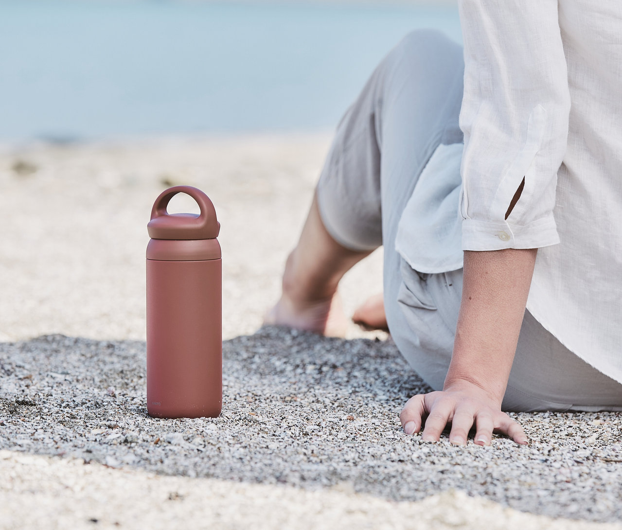 Person sitzt am Strand, daneben ein rosafarbener KINTO Tumbler »Day Off« mit Henkel auf dem Deckel.