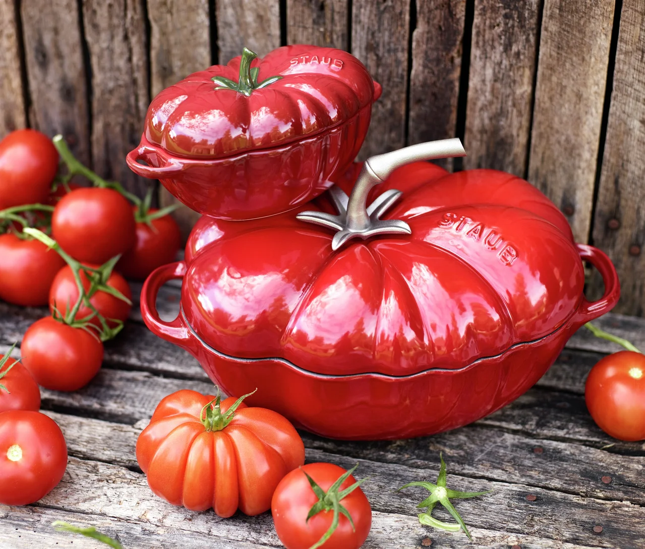 Zwei rote STAUB Gusseisen-Bräter »Tomate« auf rustikalem Holz, umgeben von frischen Tomaten. Ein kleinerer Bräter steht auf einem größeren.