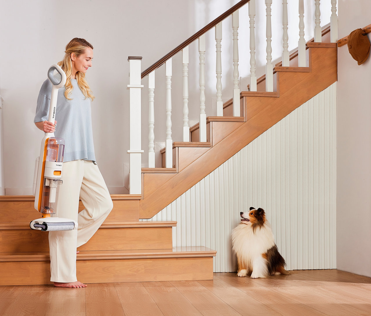 Eine Frau hält einen weißen und orangefarbenen Tineco 2-in-1-Nass-Trockensauger »Y2 Plus« und steht barfuß auf einem Holzfußboden neben einer Treppe, während ein flauschiger Hund daneben sitzt.