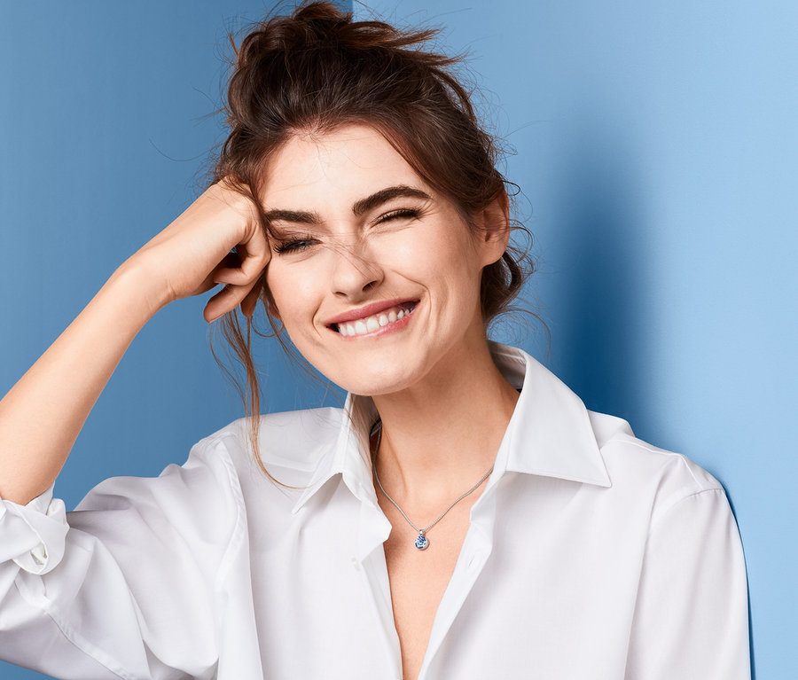 Lächelnde Frau mit braunen Haaren und weißem Hemd, den Ellbogen an die Stirn gelehnt. Sie trägt eine Kette »Solitär«, verziert mit Swarovski® Kristall.