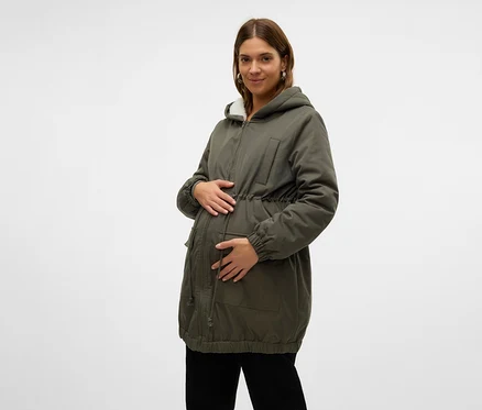 Schwangere Frau in einem grünen Parka mit weißem Kunstfell gefütterter Kapuze und Kordelzug in der Taille, die ihren Bauch hält. Sie trägt auch eine schwarze Hose.