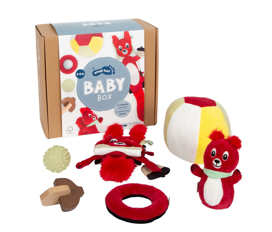 Eine braune Baby Box mit der Aufschrift "small foot BABY BOX" und mehreren Spielsachen: ein rotes Plüschtier mit Mütze, ein roter Plüschbär, ein bunter Ball, ein grüner Stachelball, ein Holzpilz und ein roter Plüschring.