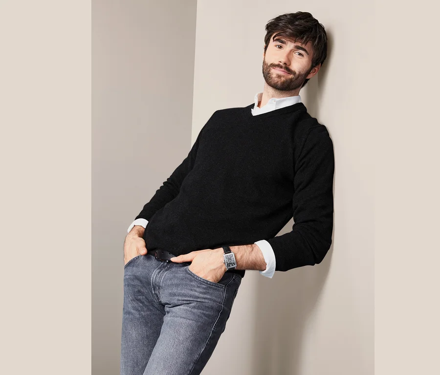 Mann lehnt an Wand, trägt Cashmere-Pullover mit V-Ausschnitt, Jeans und Uhr.