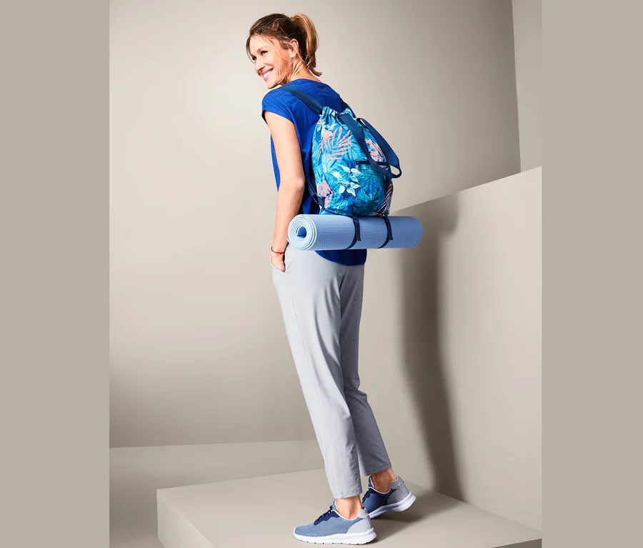 Lächelnde Frau mit blauer Yogamatte und einem 2-in-1: Rucksack und Shopper mit Blumendruck.