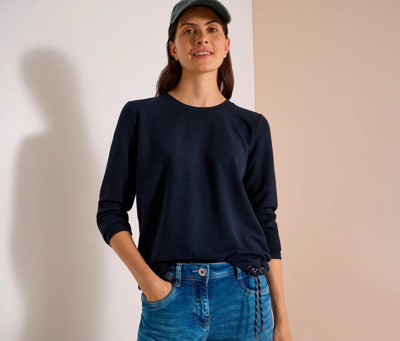 Eine lächelnde Frau trägt ein dunkelblaues, langärmeliges CECIL Shirt mit Kordelzug, Jeans und eine Kappe. Ihre Hand steckt in der Hosentasche.