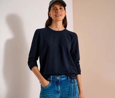 Eine lächelnde Frau trägt ein dunkelblaues, langärmeliges CECIL Shirt mit Kordelzug, Jeans und eine Kappe. Ihre Hand steckt in der Hosentasche.