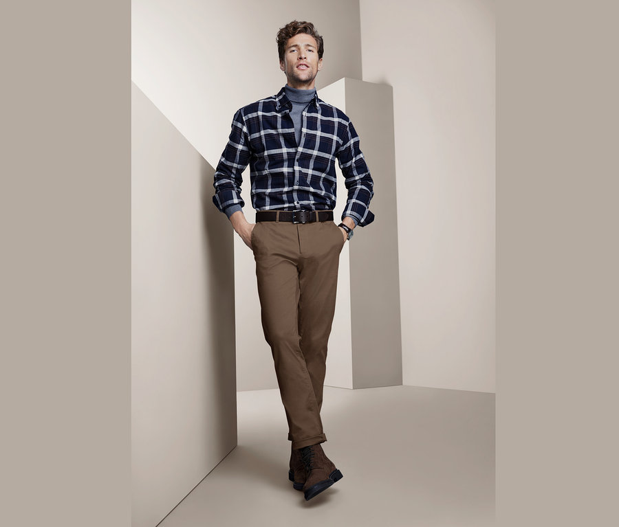 Mann posiert in braunen Chinos, kariertem Hemd und grauem Rollkragenpullover.
