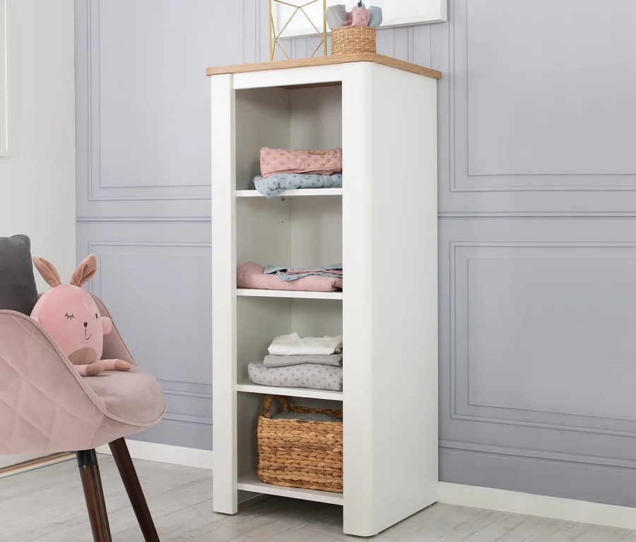Ein weißes roba Standregal "Ava" mit vier Regalböden, auf denen gefaltete Stoffe und ein Weidenkorb liegen. Daneben steht ein rosa Sessel mit einem Plüschhasen.