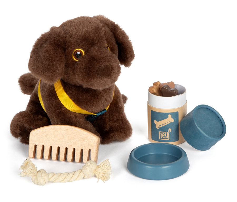 Ein brauner small foot Plüschhund mit gelbem Geschirr sitzt neben einem Kamm, einem Kauspielzeug, einem Napf und einem Behälter mit Leckerlis.