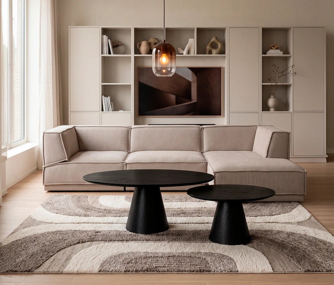 Ein modernes Wohnzimmer mit einem beigen L-förmigen Sofa, zwei runden schwarzen Couchtischen auf einem gemusterten Teppich und einer hellen WOOOD Meggy Wohnwand mit TV und Ablageschränken. Eine Pendelleuchte hängt über den Tischen, links ist ein Fenster mit Vorhängen.