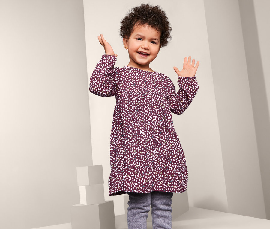 Ein lächelndes kleines Mädchen posiert in einem gemusterten Jerseykleid mit langen Ärmeln.