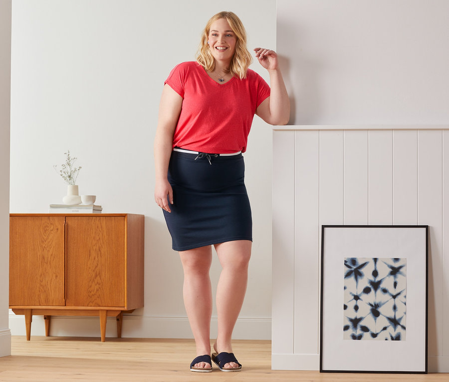 Eine Frau, die sich an eine Wand lehnt, trägt einen roten Shirt aus Leinenmix und einen Sweatrock.