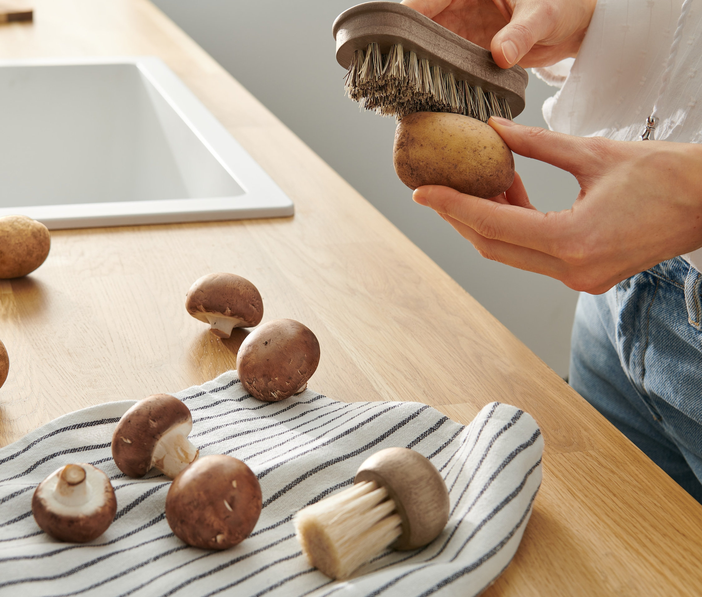 Frau putzt eine Kartoffel mit einer Gemüsebürste aus einem 2er-Set. Auf der Arbeitsfläche liegen weitere Kartoffeln und Pilze.
