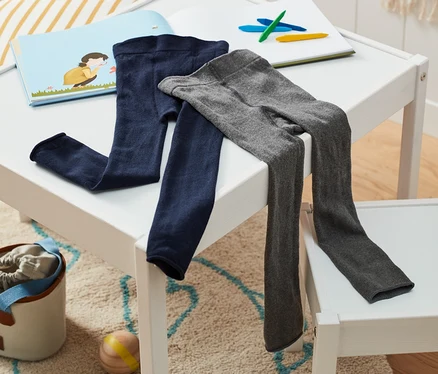 Auf einem weißen Tisch liegen blaue und graue 2 Strickleggings neben einem Buch und Buntstiften.