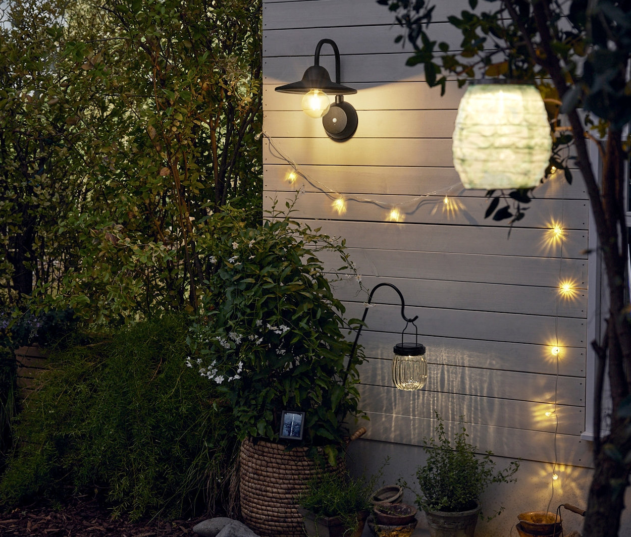 Eine LED-Akku-Wandleuchte mit zusätzlichem Wechselakku hängt an der Wand. Daneben befindet sich eine Solar-Lichterkette. Im Vordergrund befinden sich ein Solar-Lampion und eine Spieß-Solar-Leuchte.