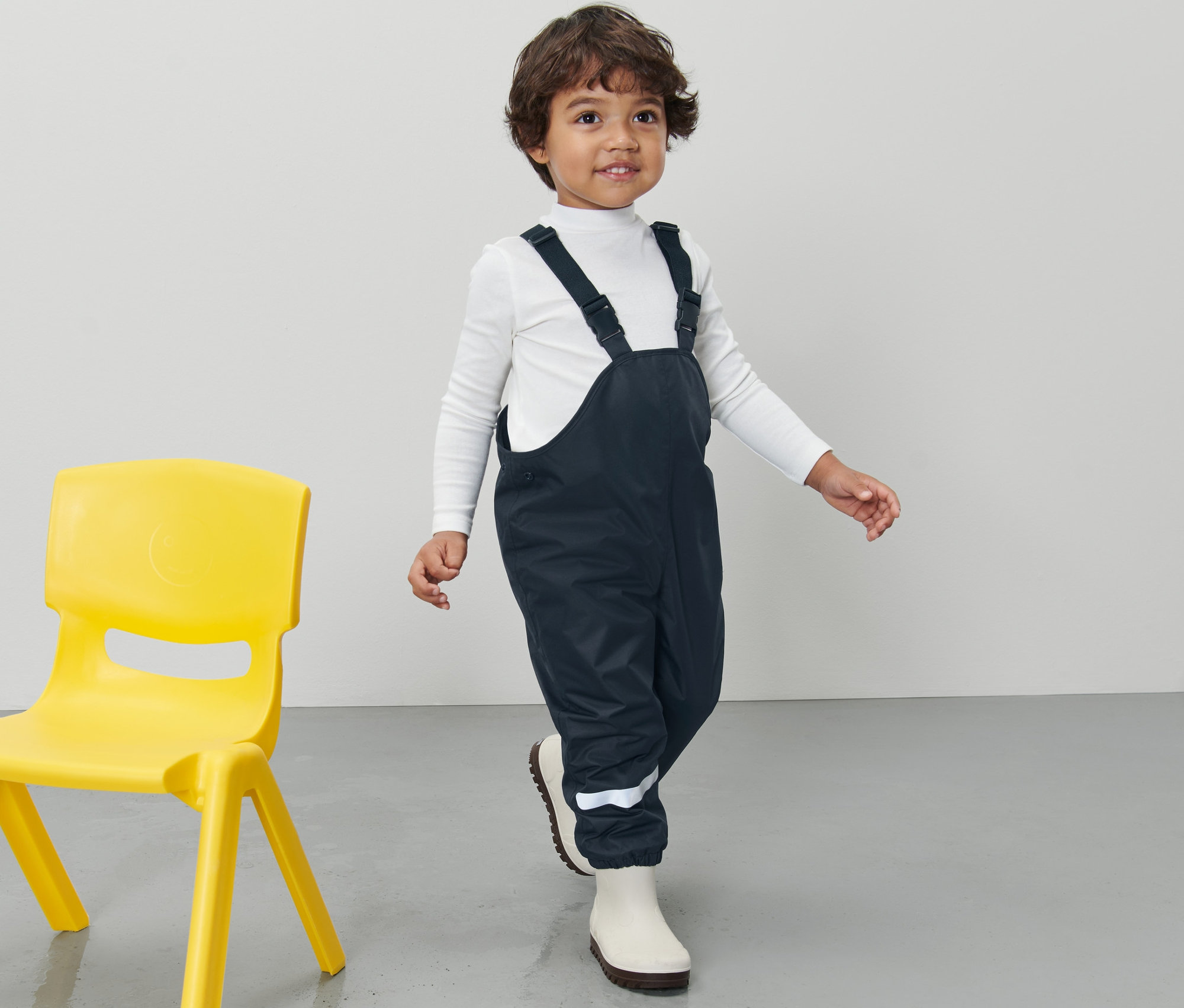 Junge trägt eine dunkelblaue Kinder-Thermo-Regenhose und einen weißen Rollkragenpullover, daneben steht ein gelber Stuhl.