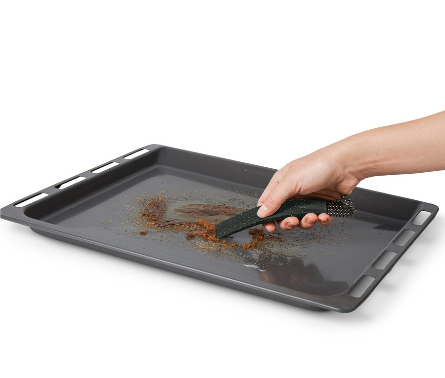 Eine Hand reinigt ein schmutziges Backblech mit einer Backofenbürste mit Schaber.