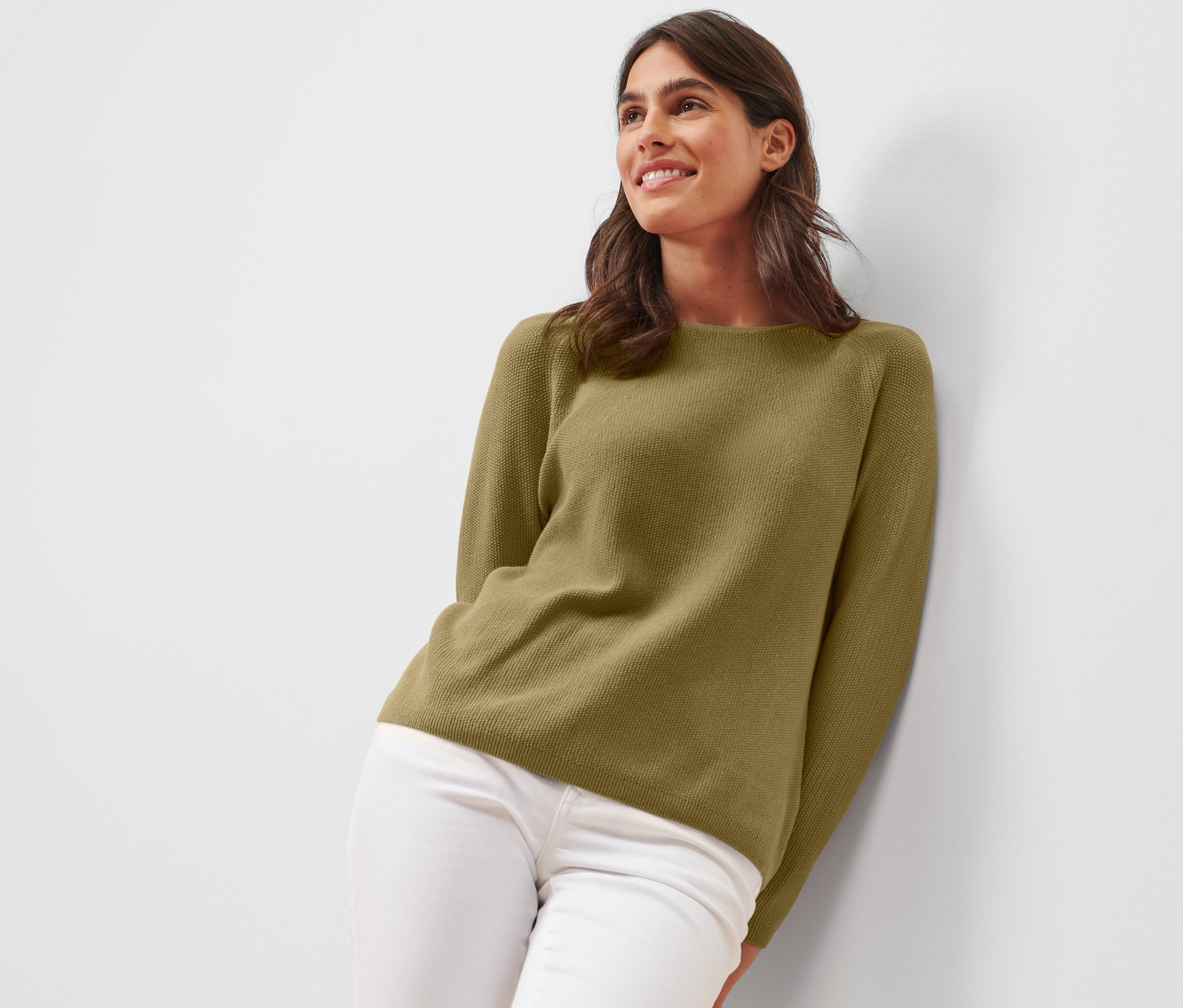 Eine Frau lehnt an einer Wand und trägt einen Strickpullover und weiße Hosen.