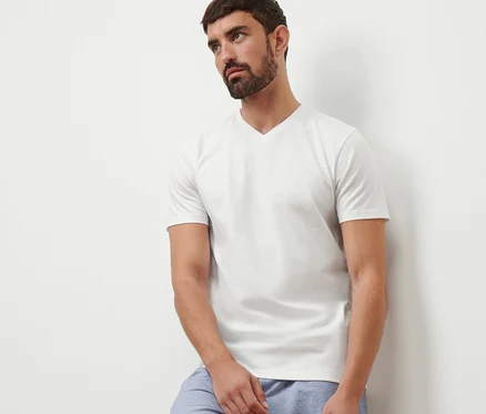 Mann lehnt an einer Wand, trägt ein weißes V-Ausschnitt T-Shirt und blaue Shorts.