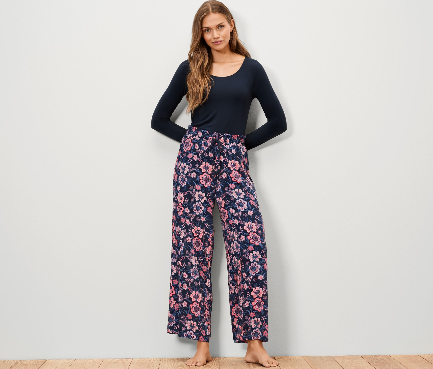 Eine Frau steht und trägt eine Web-Relaxhose mit Blumenmuster und ein blaues Langarmshirt.