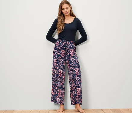 Eine Frau steht und trägt eine Web-Relaxhose mit Blumenmuster und ein blaues Langarmshirt.