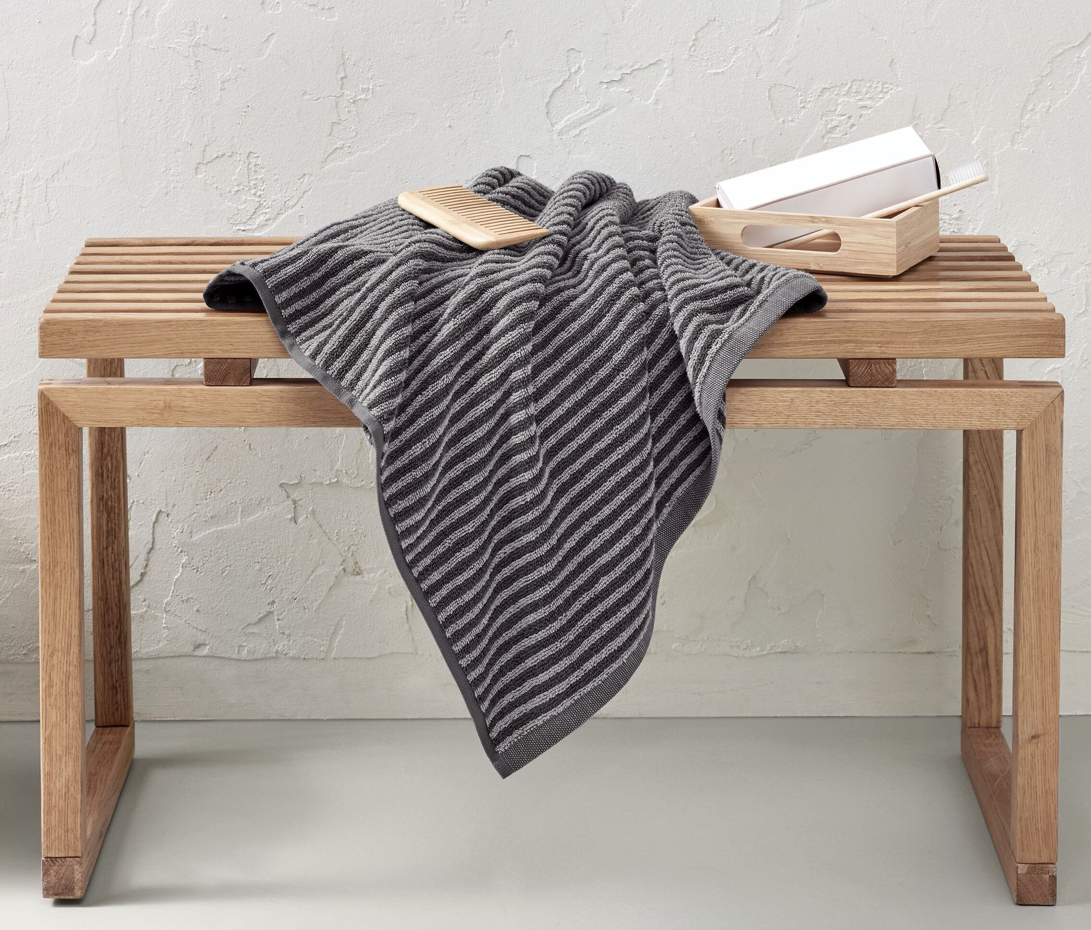 Auf einer Holzbank liegt ein hochwertiges, graues Badetuch. Neben der Bank steht ein Weidenkorb.