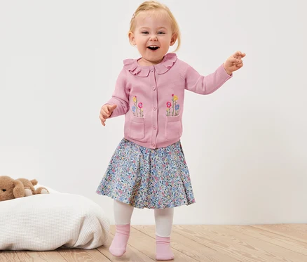 Lächelndes Kleinkind winkt mit den Händen und trägt eine rosa Baby-Strickjacke mit Blumen, einen geblümten Rock und rosa Socken.