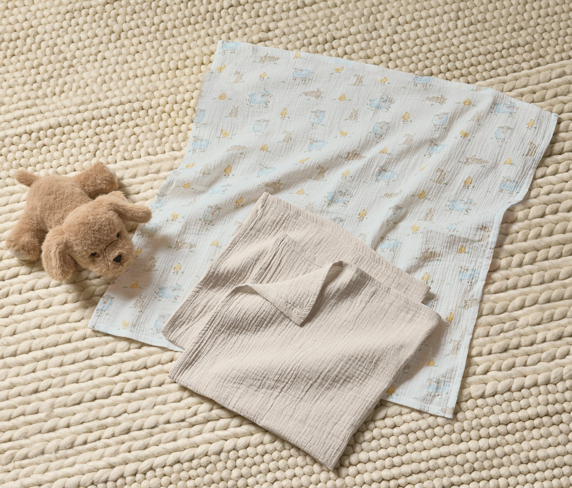 Ein Stofftier-Hund und 3 Baby-Musselin-Tücher auf einem gestrickten Teppich.