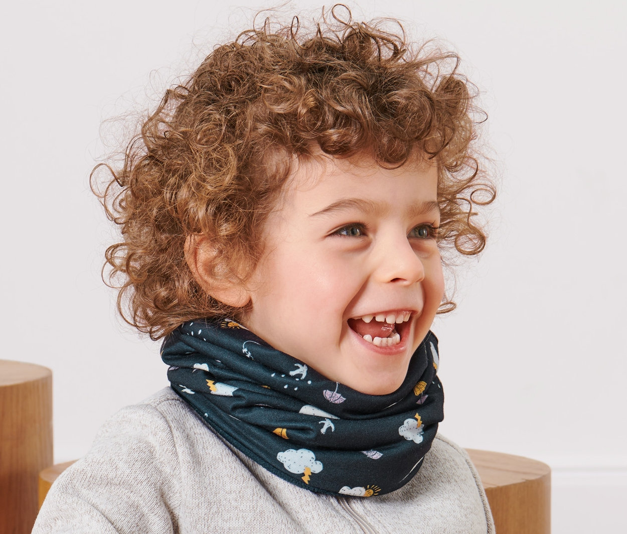 Lächelnder kleiner Junge mit lockigem Haar sitzt und trägt ein Kleinkind-Multifunktionstuch.