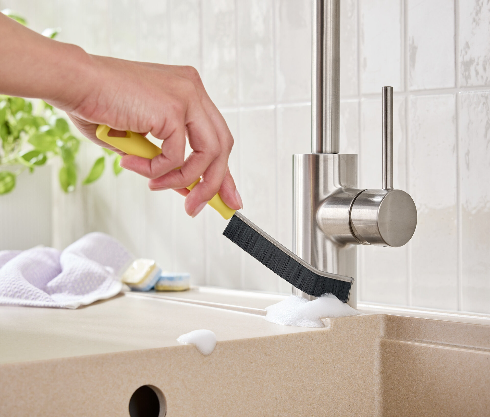 Eine Hand reinigt einen Wasserhahn mit einer gelben Bürste in einem Waschbecken.