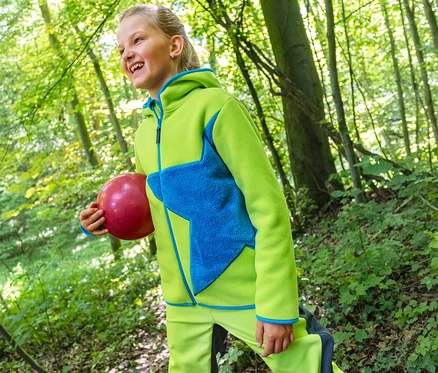 Lächelndes Mädchen in einer grünen Jako-O Kuschelfleecejacke mit blauem Stern und passender Hose, das einen roten Ball im Wald hält.