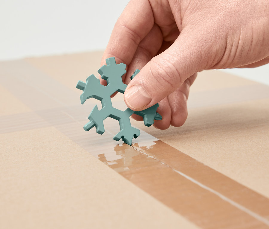 Eine Hand hält einen schneeflockenförmigen Multifunktionsschlüssel und schneidet damit Klebeband auf einem Karton.