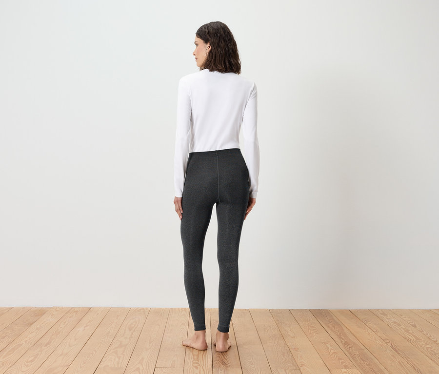 Frau steht mit dem Rücken zur Kamera und trägt ein weißes Langarmshirt und graue Leggings.