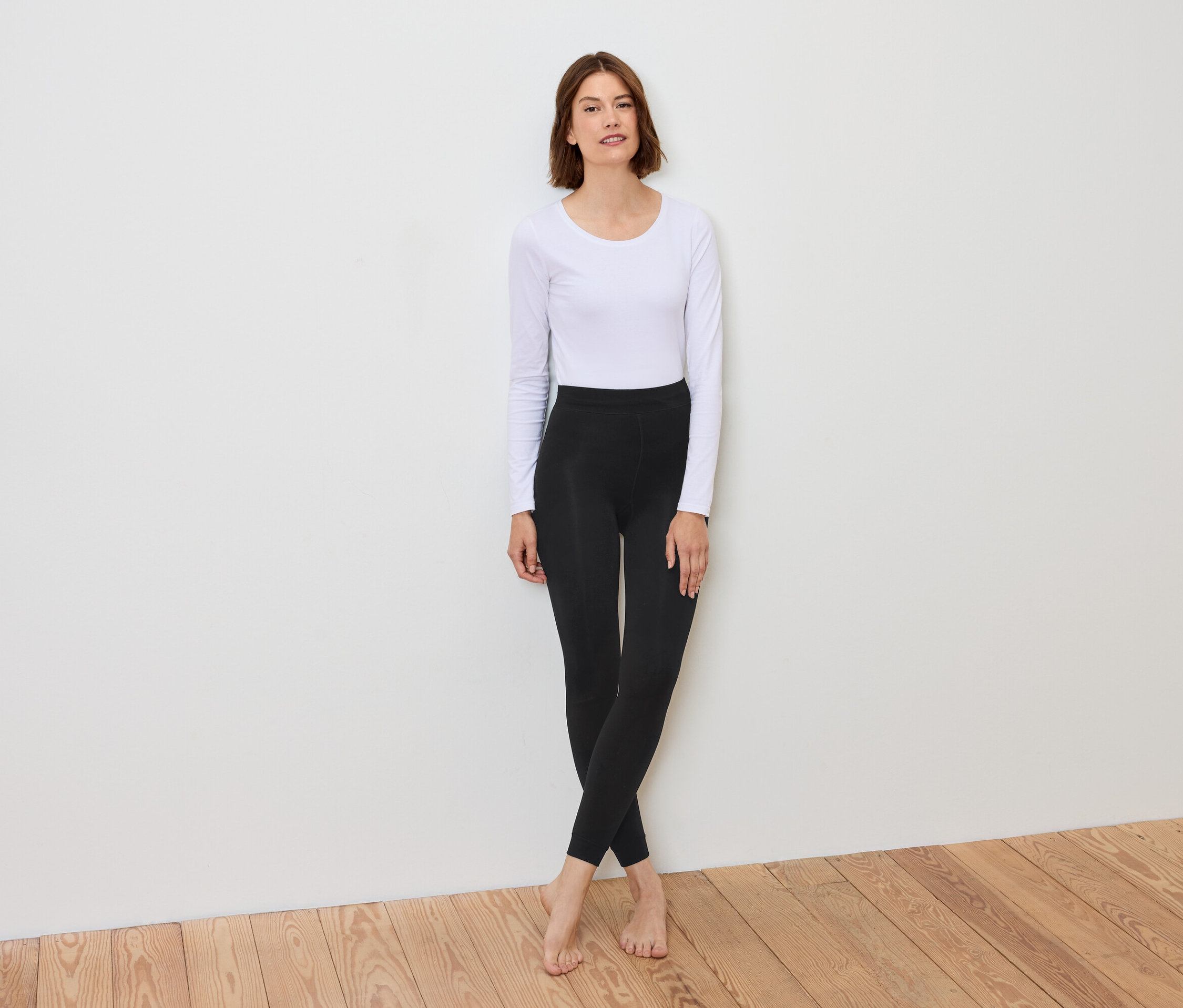 Eine Frau steht vor einer weißen Wand und trägt ein weißes Langarmshirt und schwarze Leggings.