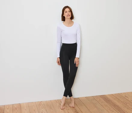 Eine Frau steht vor einer weißen Wand und trägt ein weißes Langarmshirt und schwarze Leggings.
