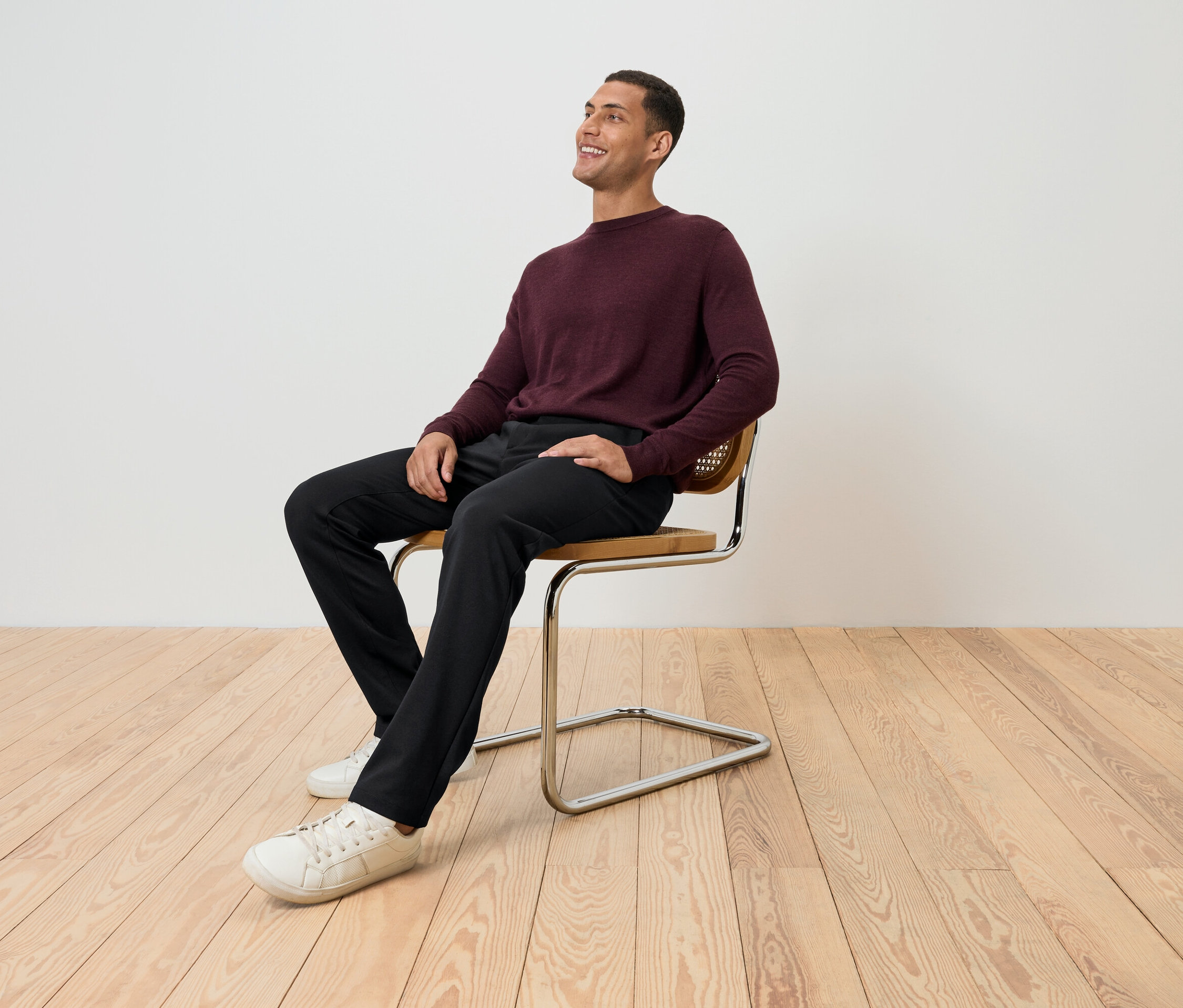 Ein Mann sitzt auf einem Stuhl in einem bordeauxfarbenen Pullover, schwarzen Hosen und weißen Schuhen und schaut nach oben.