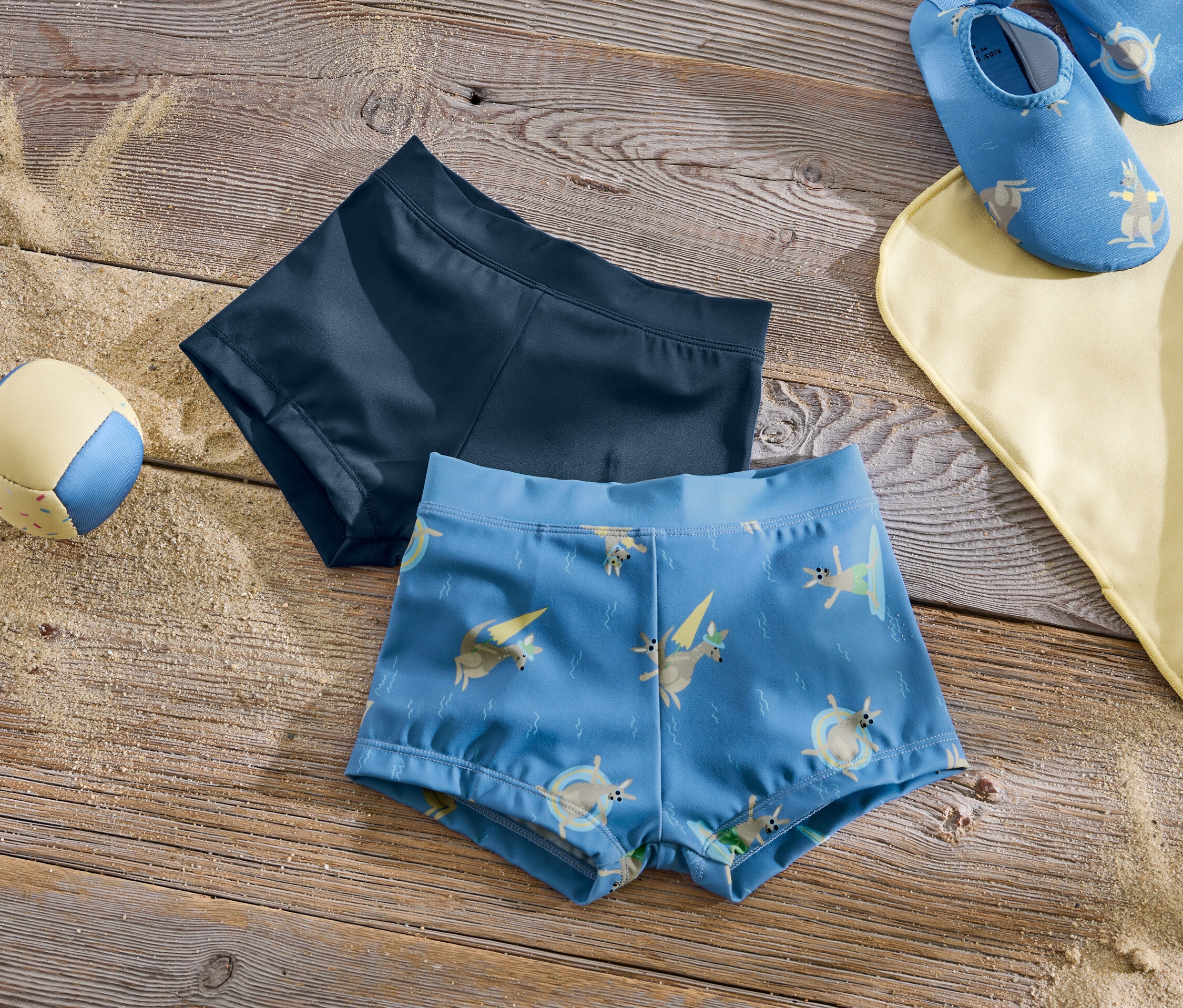 Zwei Kinder-Badehosen liegen neben einem Ball und einem Badetuch auf einem Holztisch.