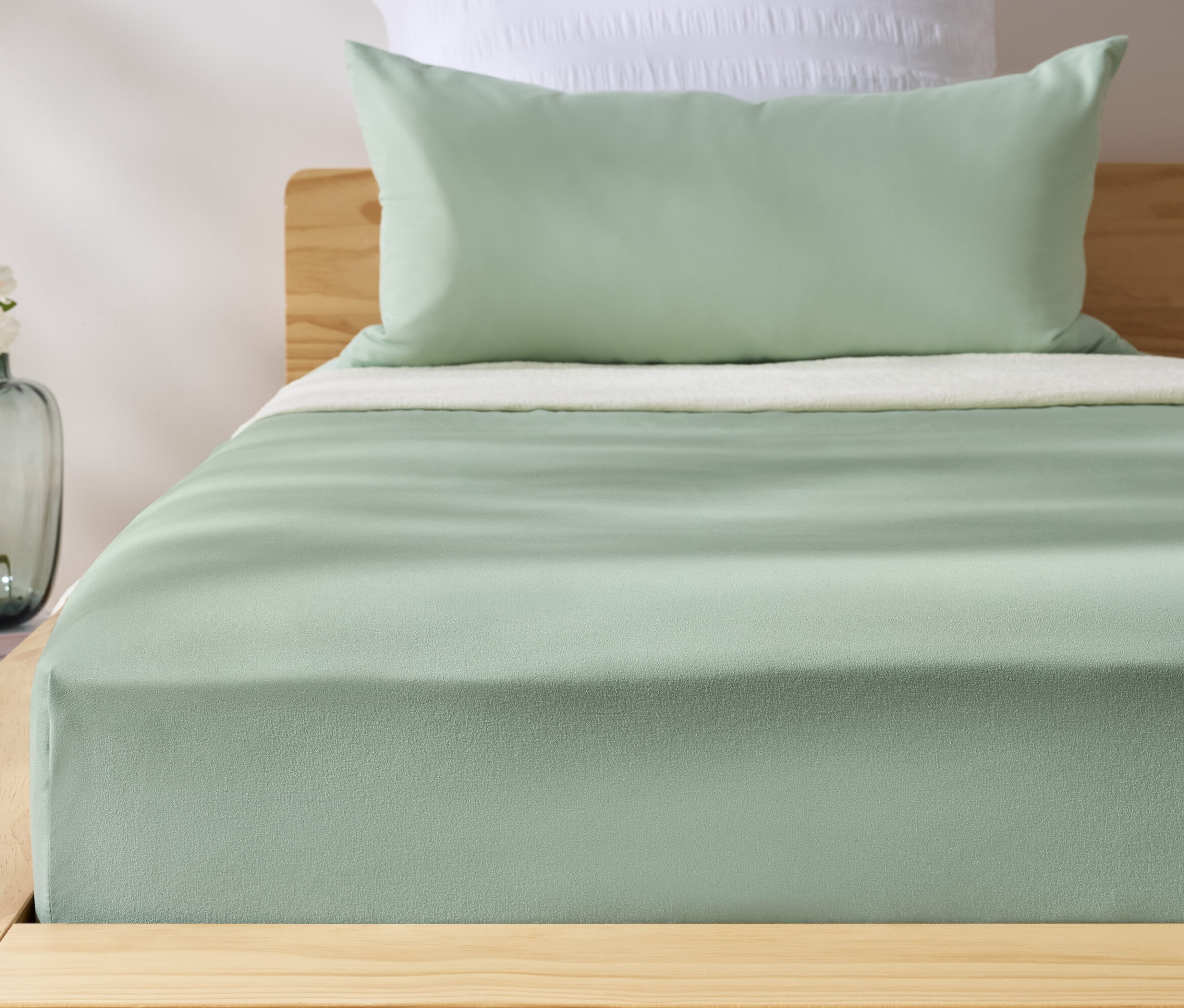 Grünes Microfaser-Spannbettlaken auf einem Bett. Neben dem Bett steht eine Vase mit Blumen.