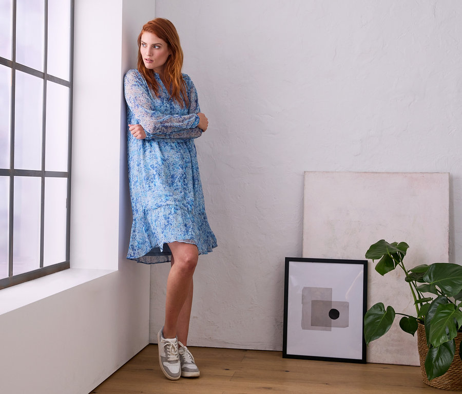 Frau im Kleid mit Alloverprint steht am Fenster.