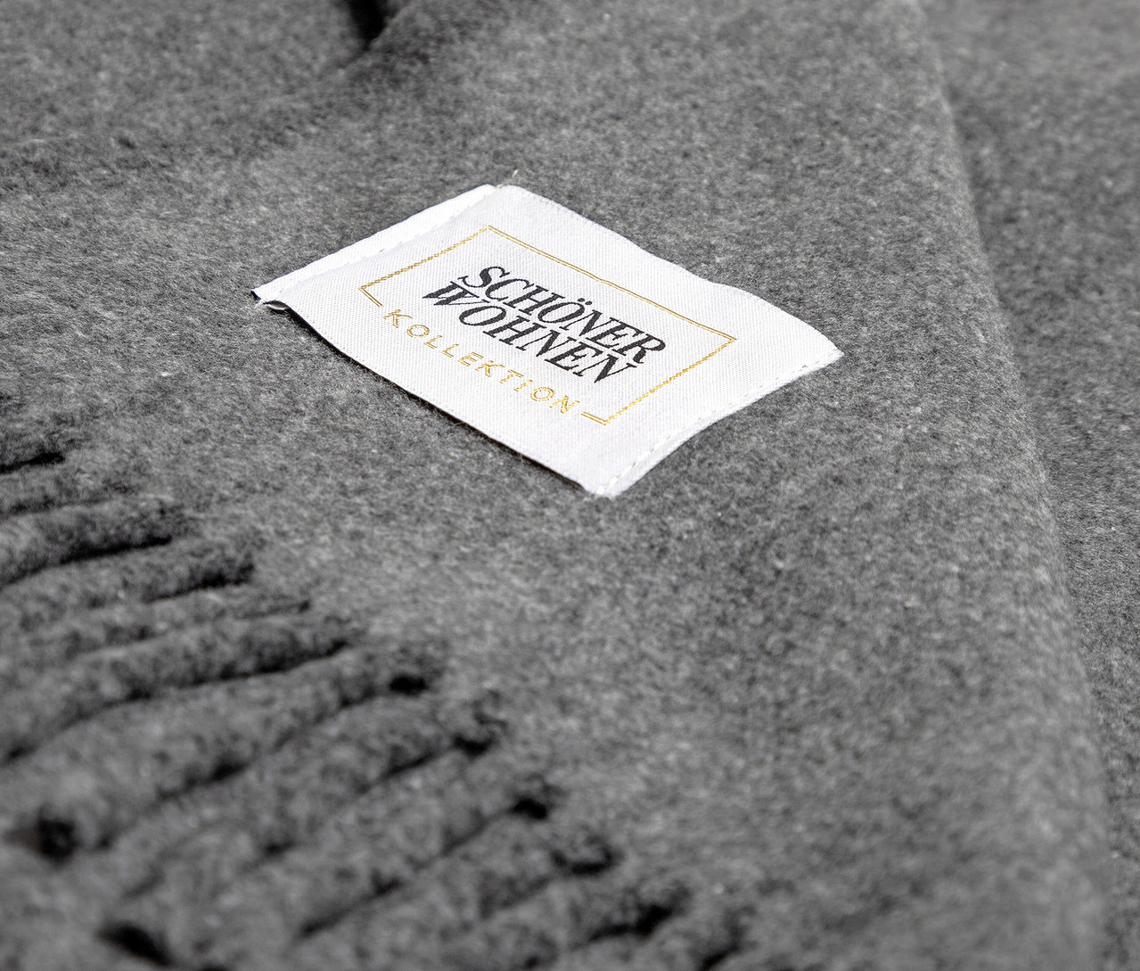 Detail einer grauen Decke mit Fransen und dem Etikett "SCHÖNER WOHNEN - KOLLEKTION".