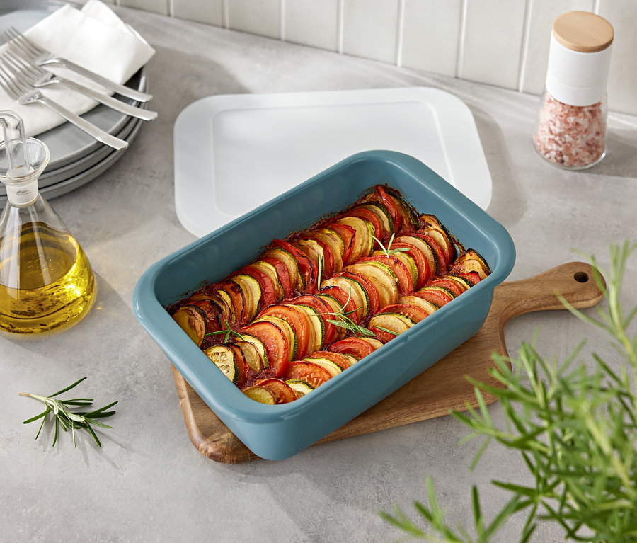 Gemüse in blauer Auflaufform auf Holzbrett. Daneben Ölflasche und Gewürzstreuer.