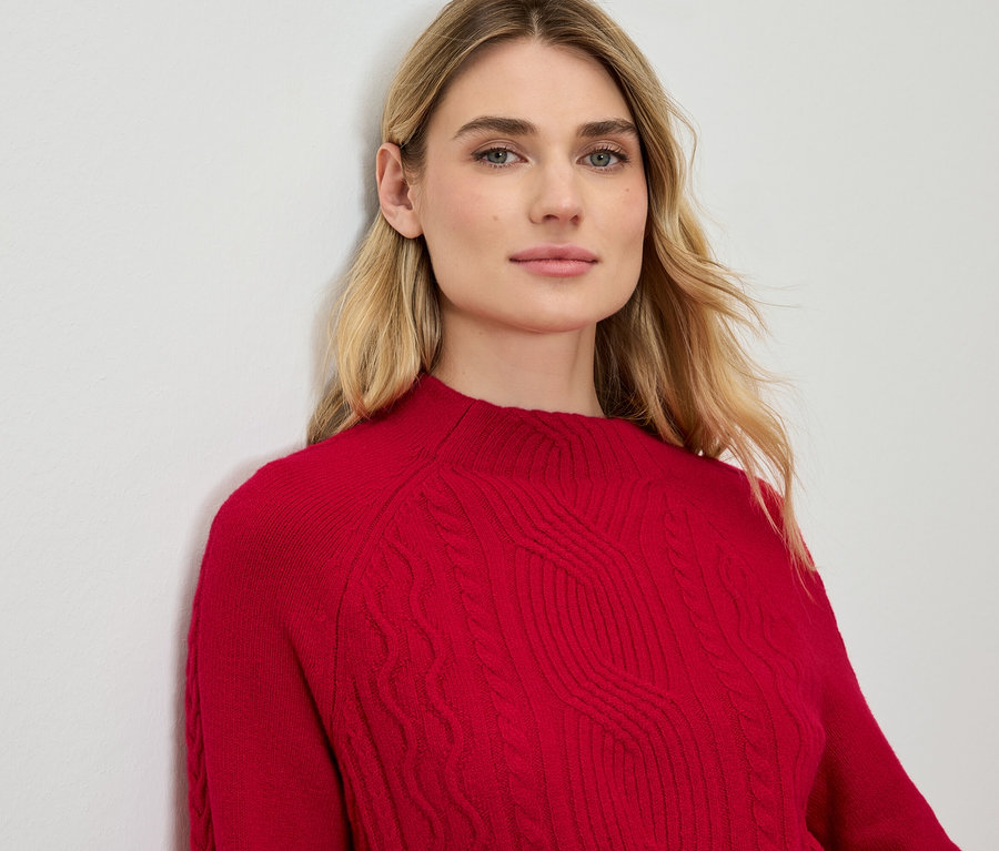 Blonde Frau lehnt an einer Wand und trägt einen roten Pullover.