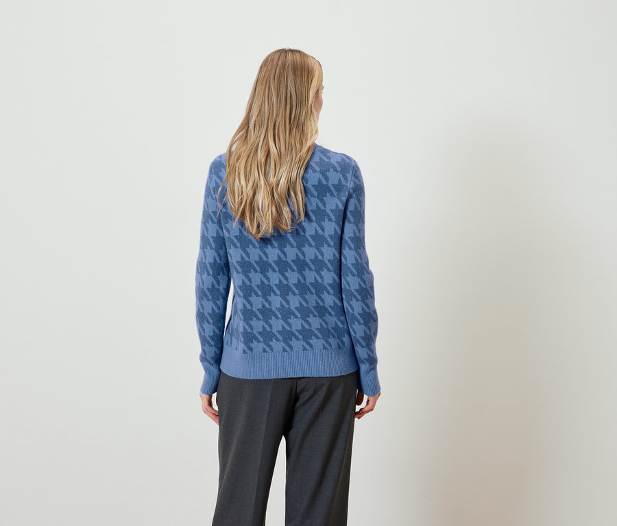 Eine Frau mit blonden Haaren, von hinten gesehen, trägt einen blauen Hahnentrittpullover und graue Hosen.