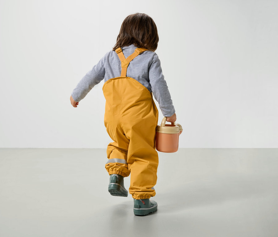 Junge in gelber Latzhose und Gummistiefeln geht über den Boden und trägt eine rosa Gießkanne.