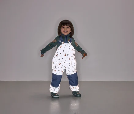 Ein lächelndes Kind steht mit ausgestreckten Armen und trägt eine Latzhose mit Tiermuster und marineblauen Hosenbeinen.