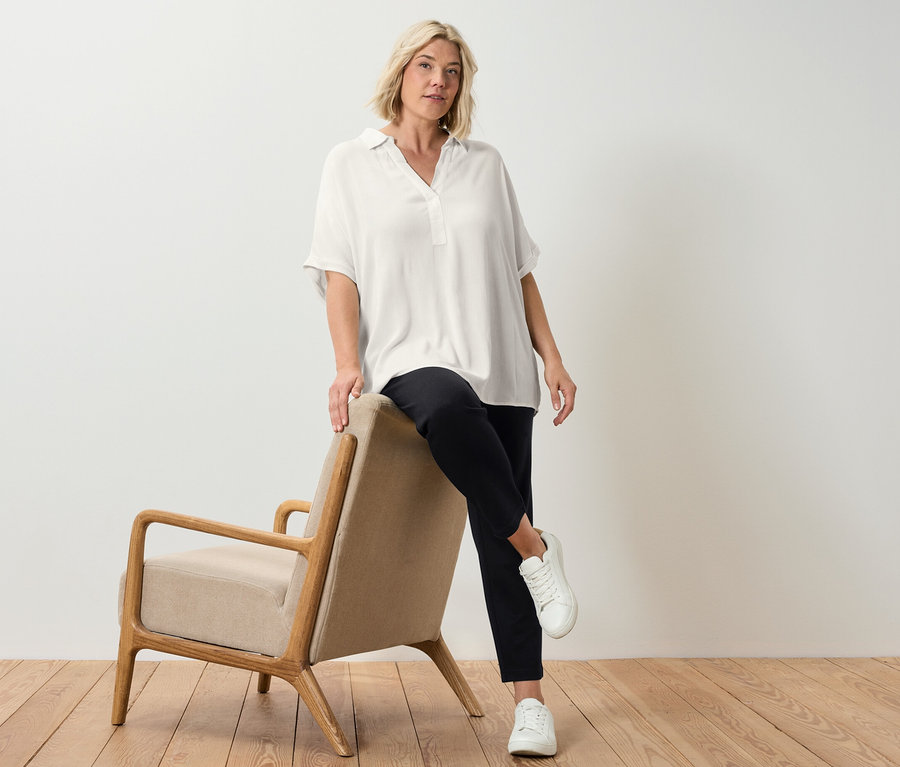 Blonde Frau posiert an einem beige-farbenen Sessel, sie trägt eine cremeweiße Bluse.