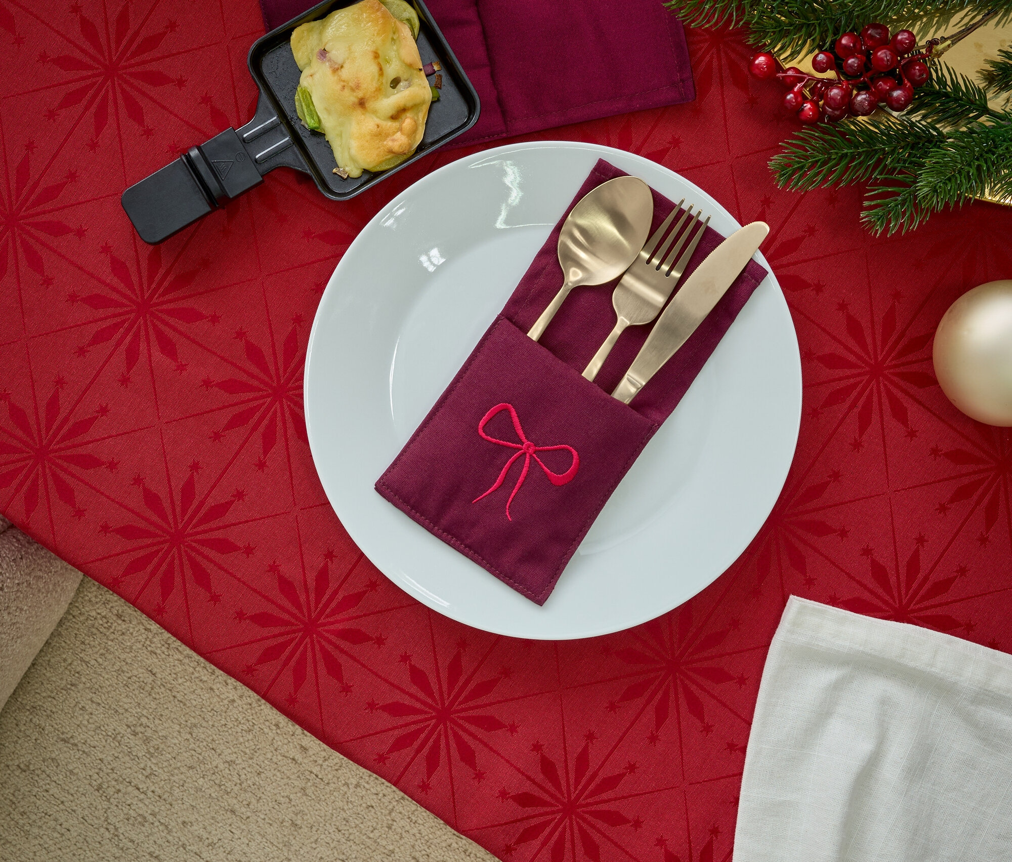 Teller mit besticktem Stoffserviettenhalter, goldenes Besteck, Raclette-Pfännchen und Weihnachtsdekoration auf roter Tischdecke.
